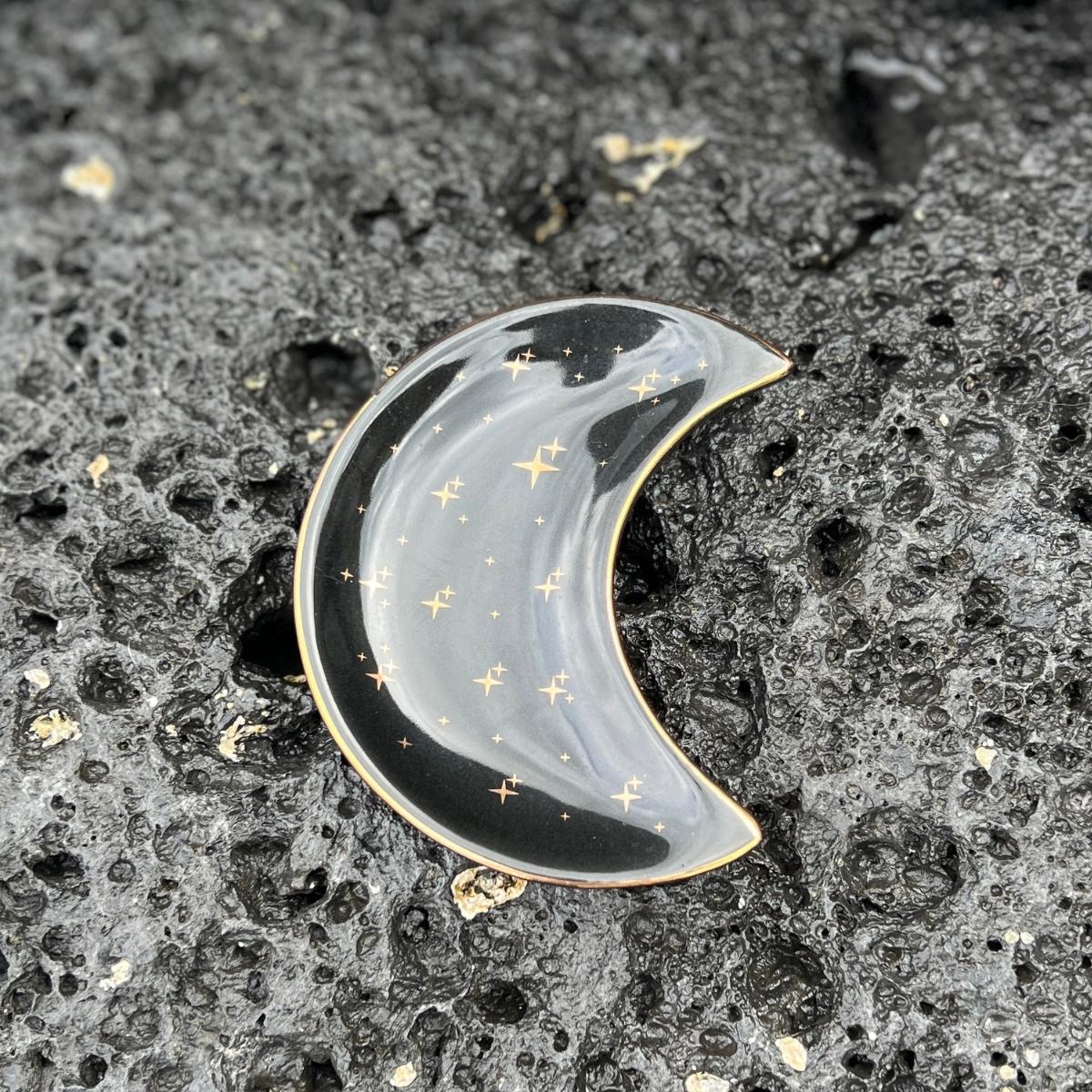 The Celeste Jewelry Dish is a beautiful and ethereal crescent-shaped ceramic dish, perfect for storing and displaying your favorite jewelry pieces. The dish features a stunning cosmic-inspired design, with gold stars and a black background that create a celestial and magical look.
