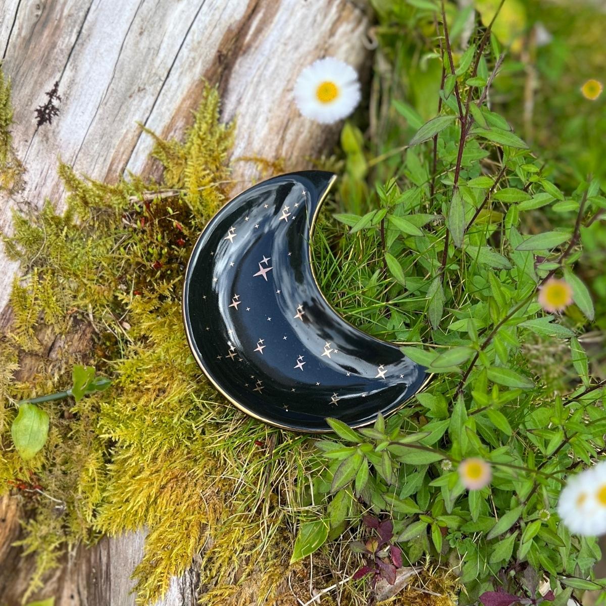 The Celeste Jewelry Dish is a beautiful and ethereal crescent-shaped ceramic dish, perfect for storing and displaying your favorite jewelry pieces. The dish features a stunning cosmic-inspired design, with gold stars and a black background that create a celestial and magical look.